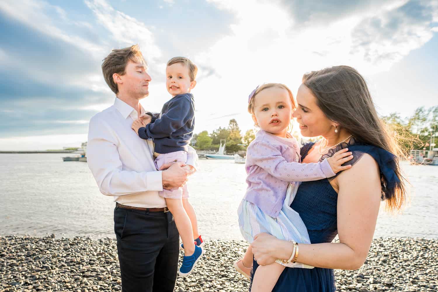 A couple on a pebble beach, each holding a child. The man holds a young boy and the woman holds a young girl. The cloudy sky and water is in the background.