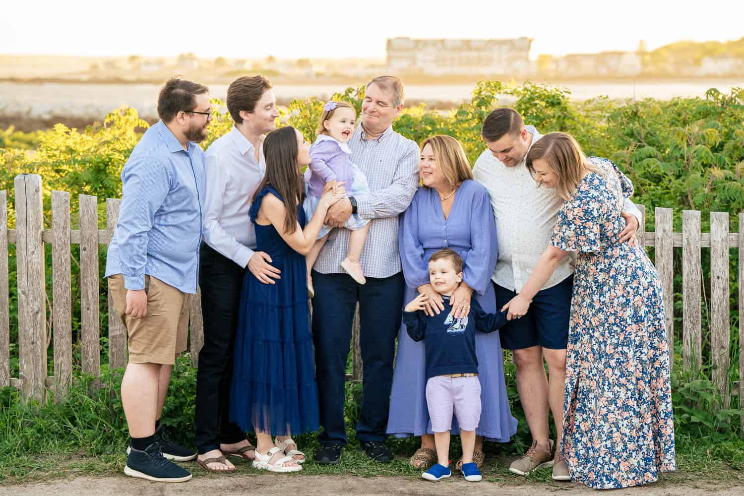 A group of eight adults and two children stand together in front of a wooden fence and greenery, smiling and interacting with each other.