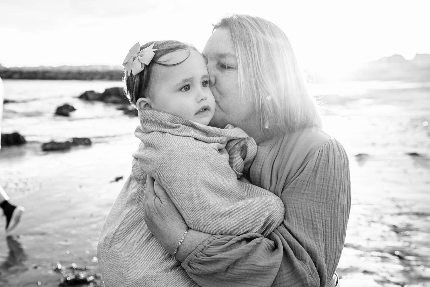 A woman lovingly kisses a small child wrapped in a blanket while standing on a beach. The background features a bright sky and the ocean.