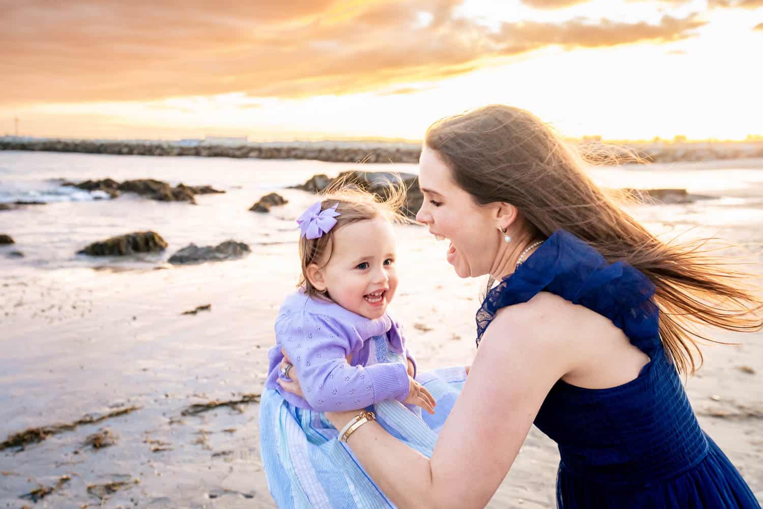 A woman and a young child smile and play together on a beach during sunset. The woman holds the child, who is wearing a purple dress with a matching bow in her hair.