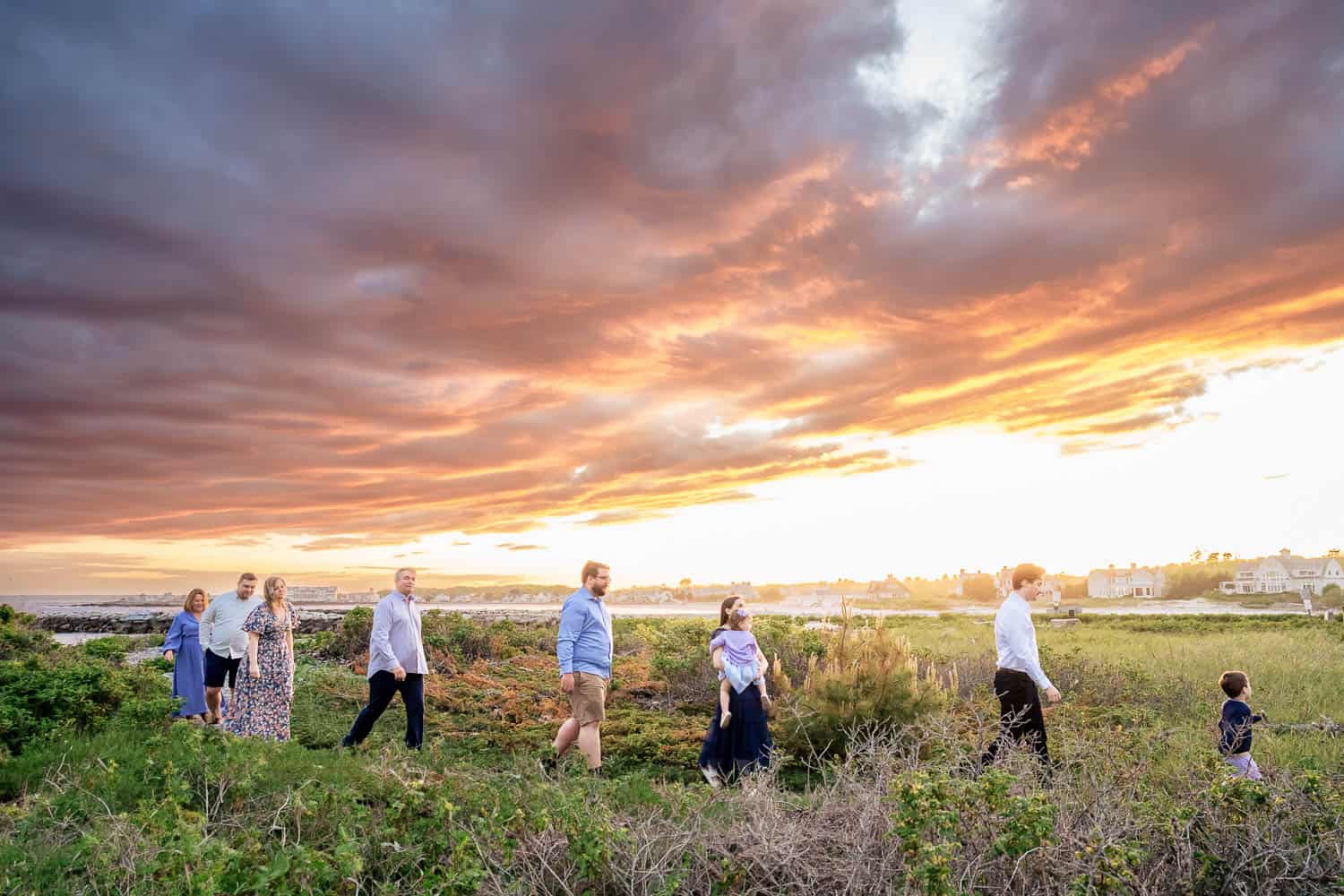 Family Photography 26 A group of seven people walks through a grassy field with a colorful sunset and cloud-filled sky in the background.