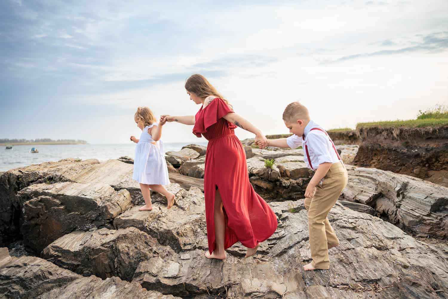 Two children and their mother holding hands and walking on rocky terrain by the sea. The mother is in a red dress in the middle.