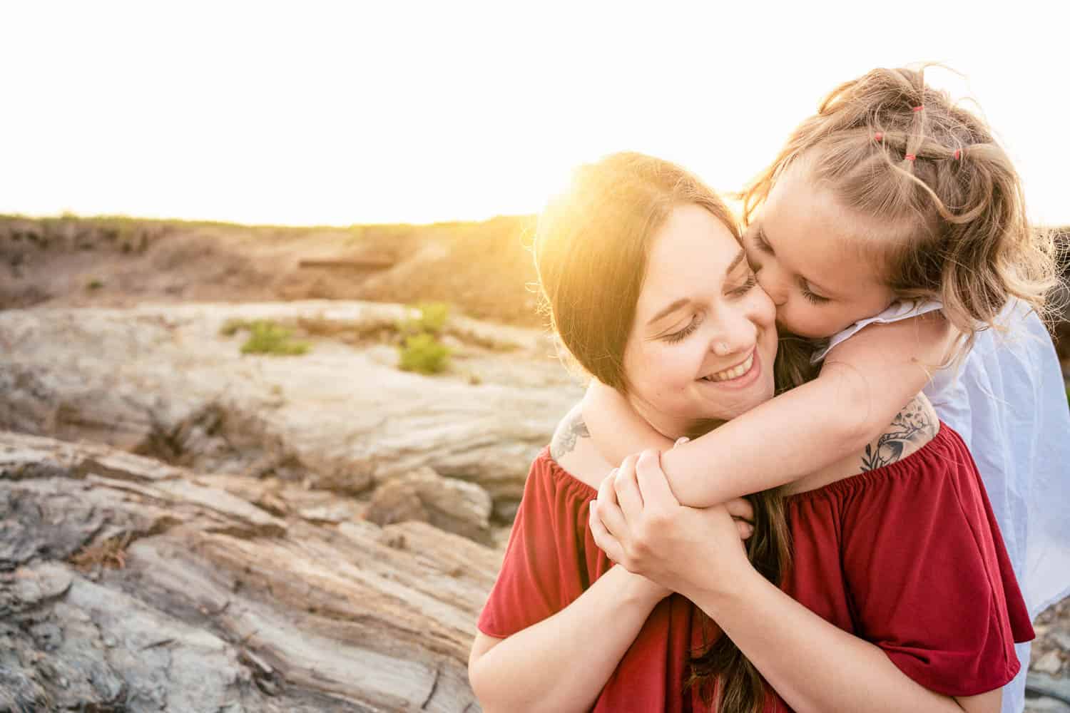 A child hugs and kisses a smiling woman from behind at a rocky outdoor location with sunlight in the background.