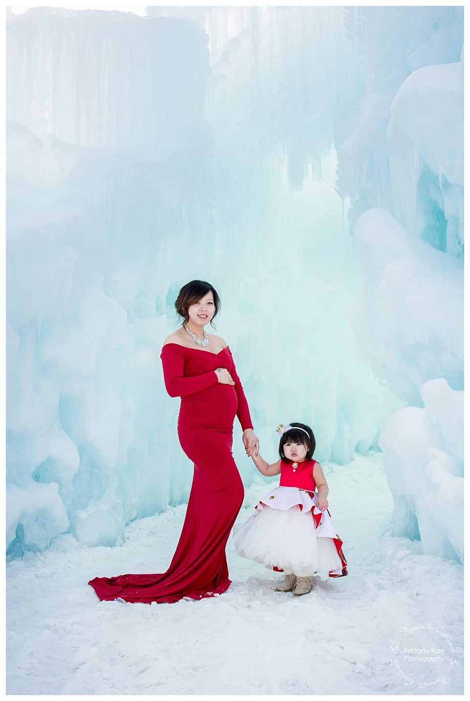 Woman and daughter post in red dresses for maternity portraits at the Ice Castles in New Hampshire